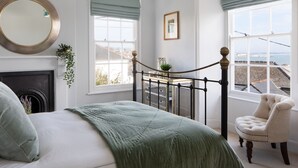 A bedroom with a bed, a fireplace, a view of the sea, and a chair by the window. - Belmont (Penzance)