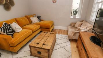 A living room with a mustard sofa, a beige armchair, a wooden coffee table, and a TV on a wooden stand.