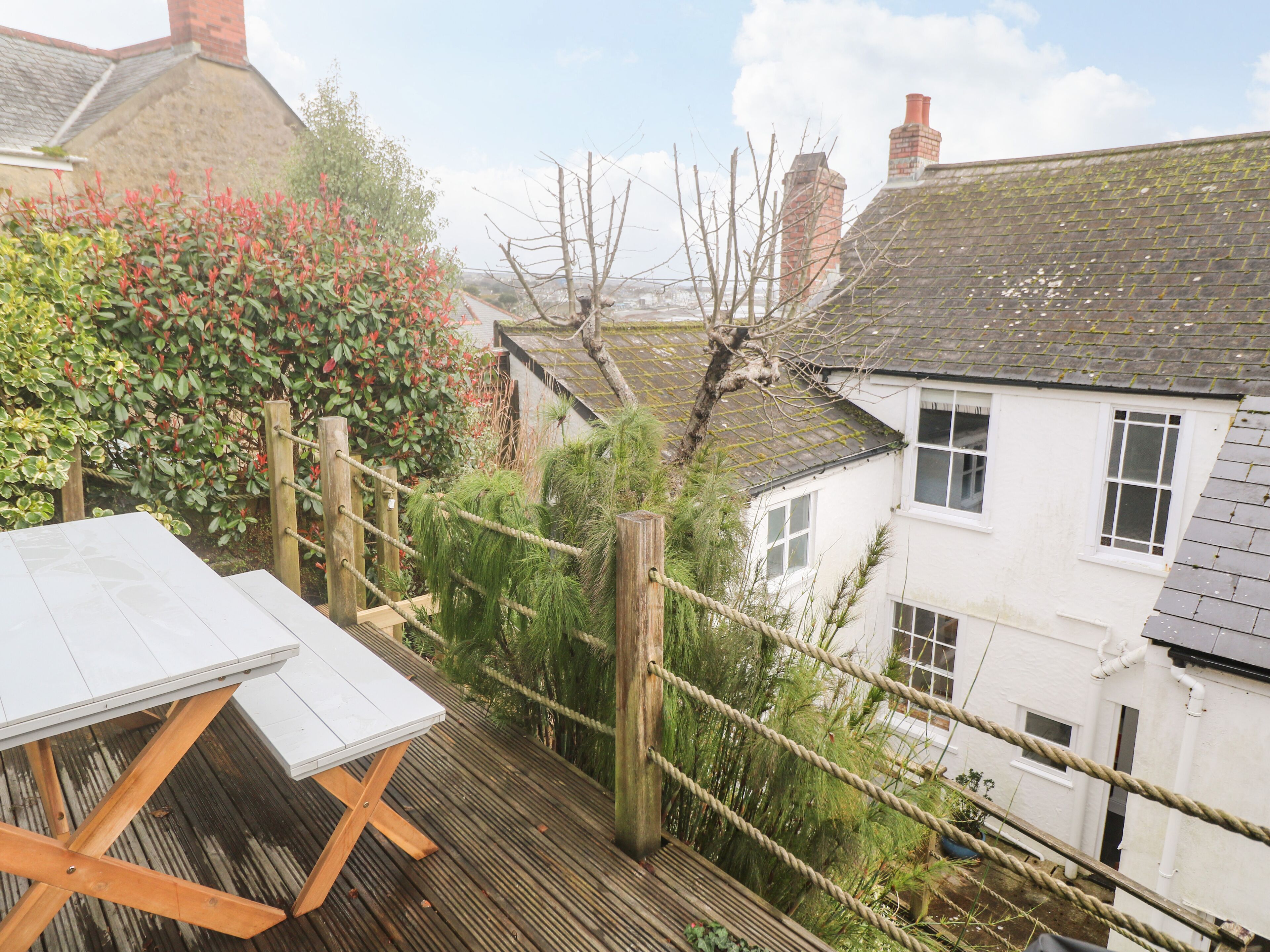 A wooden deck with a table, surrounded by lush greenery and a view of a white house with a sloped roof.