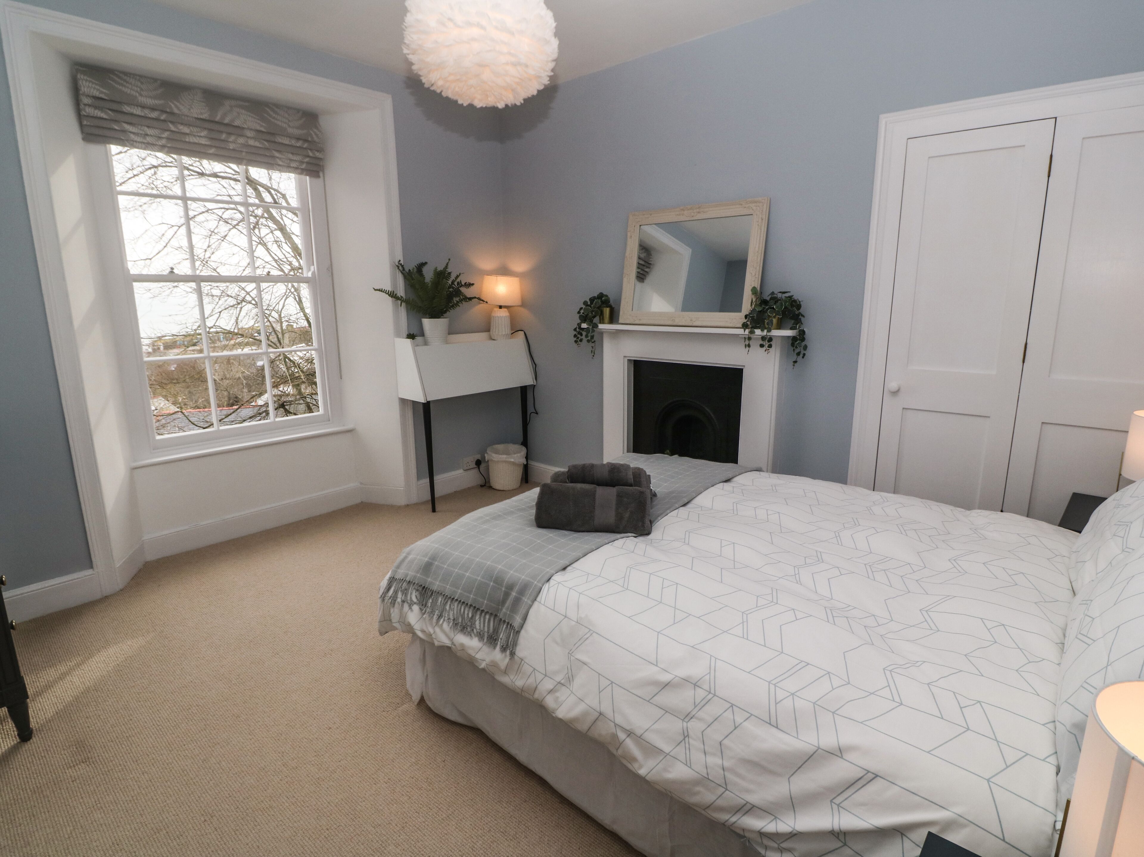A bedroom with a bed, a bedside table, a lamp, a mirror, a fireplace, and a window with a view of trees.