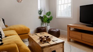 A living room with a yellow sofa, a wooden coffee table with blocks, a TV on a wooden stand, and a potted plant. - Belmont (Penzance)