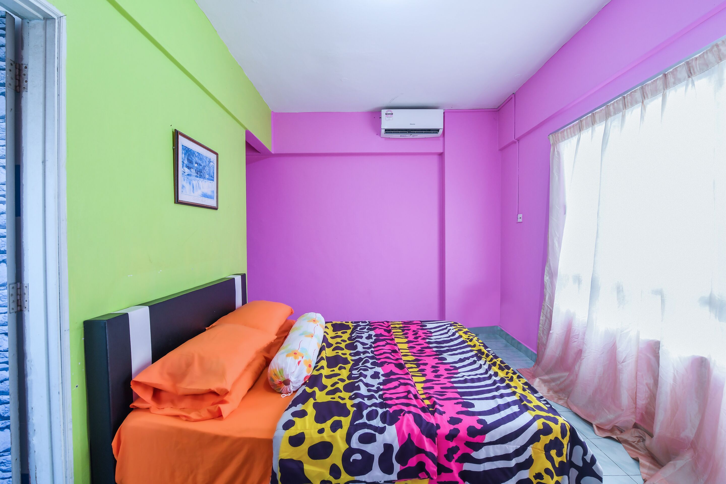 A room with a bed, a window with sheer curtains, a picture on the wall, and an air conditioning unit.