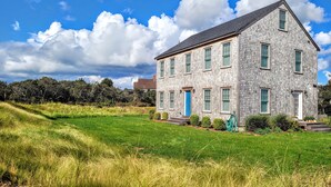 A two-story stone house with a blue door and green shutters, surrounded by a grassy field and trees under a partly cloudy sky. - Beautiful 4 bed with Pool + Spa (Nantucket)