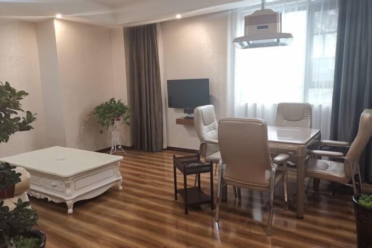 A modern living room with a wooden floor, a white sofa, a dining table with chairs, a TV, and a large window with curtains.