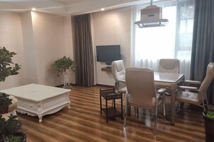 Una sala de estar moderna con piso de madera, un sofá blanco, una mesa de comedor con sillas, un televisor y un ventanal con cortinas.