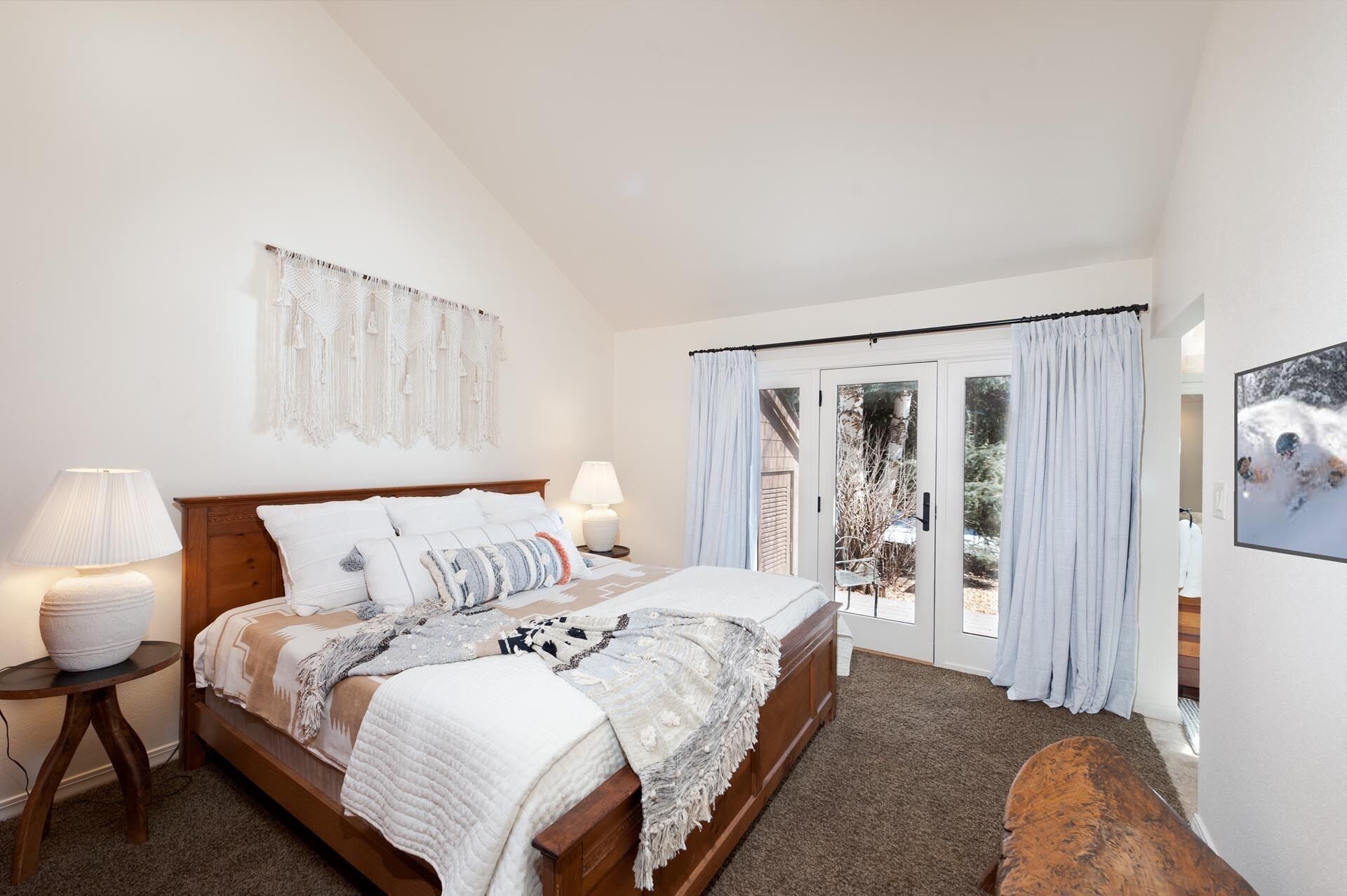 A bedroom with a large bed, two bedside lamps, a macrame wall hanging, a sliding glass door to a patio, and a framed picture on the wall.