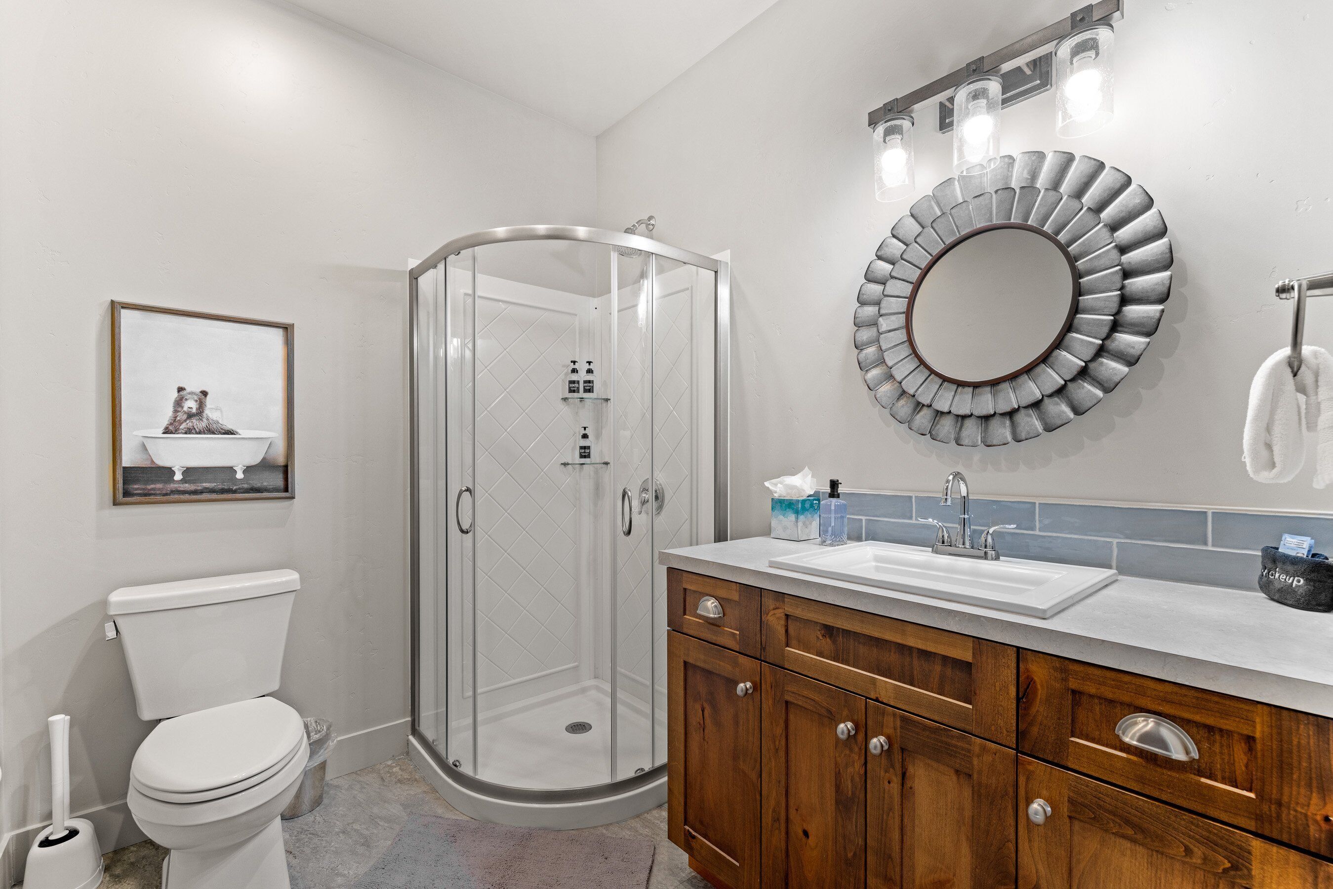Un baño con cabina de ducha de vidrio, inodoro, lavabo con tocador de madera y espejo redondo.