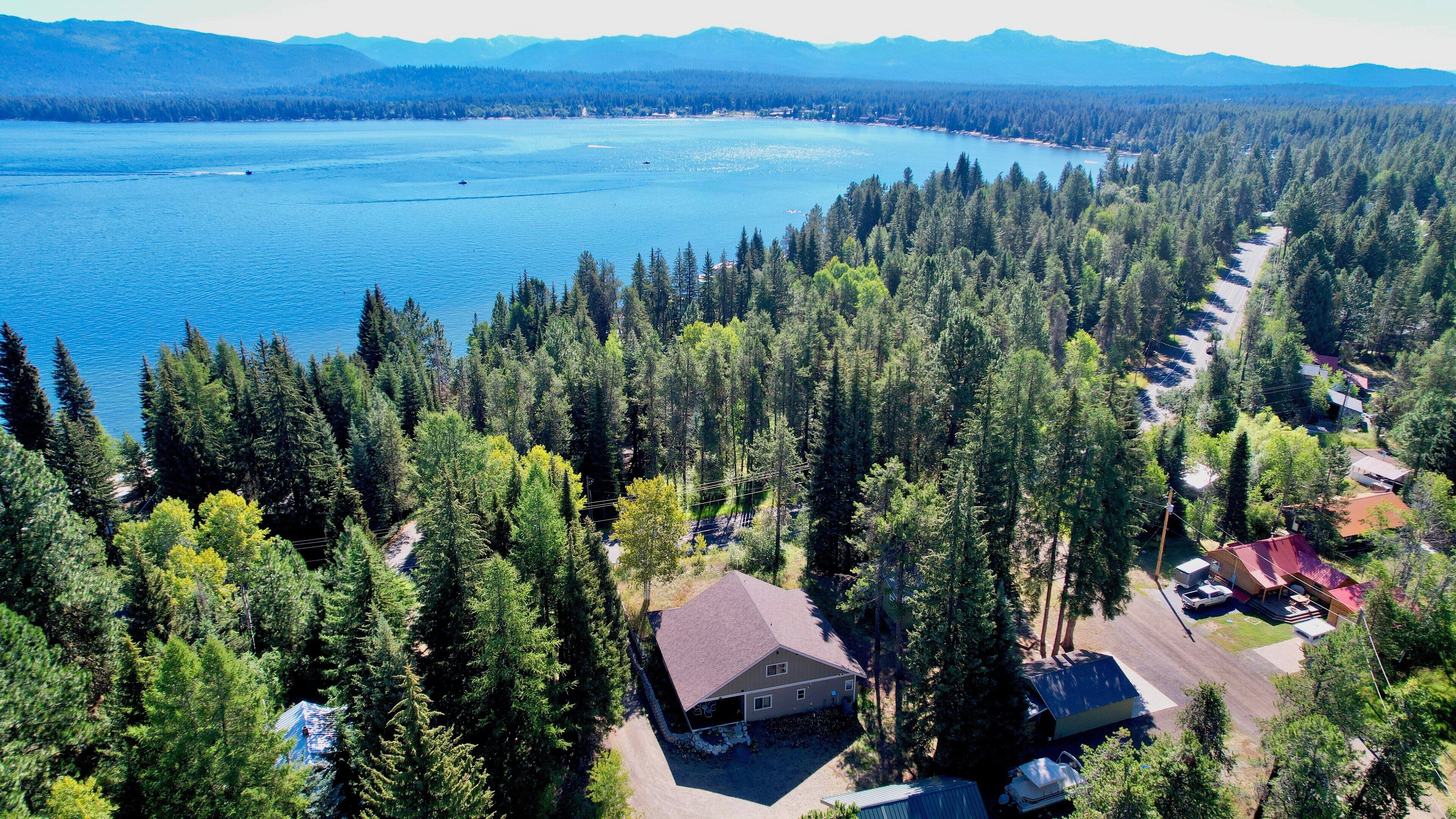 An aerial view of a forested area with a large body of water, a road, and a house with a covered porch.
