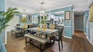 A dining area with a long table, benches, and chairs. There is a kitchen in the background with cabinets and a microwave.