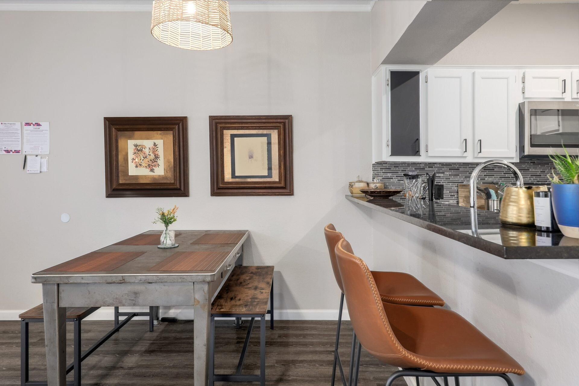 A modern kitchen with white cabinets, a stainless steel sink, and a built-in microwave. There is a wooden dining table with a bench and a brown leather chair. Two framed artworks are hung on the wall above the table.