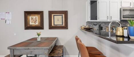 A modern kitchen with white cabinets, a stainless steel sink, and a built-in microwave. There is a wooden dining table with a bench and a brown leather chair. Two framed artworks are hung on the wall above the table.