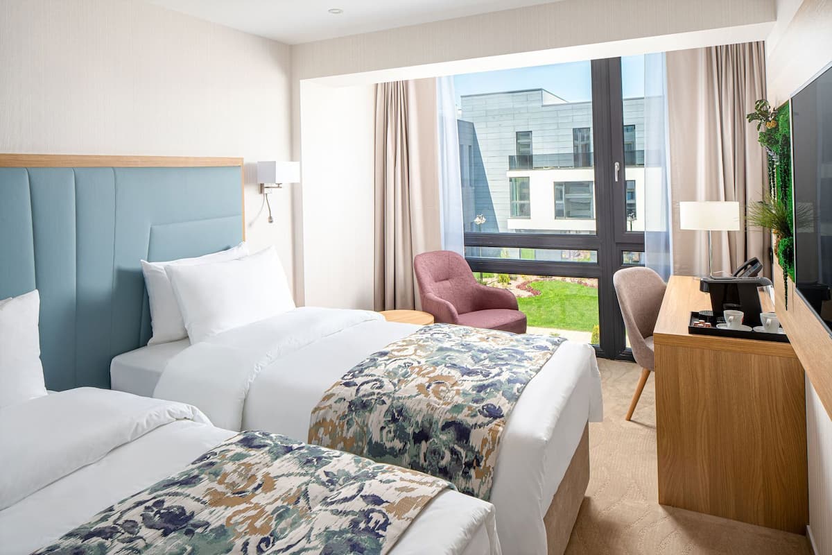 A hotel room with two beds, a desk with a telephone, a chair, and a large window with a view of a building.