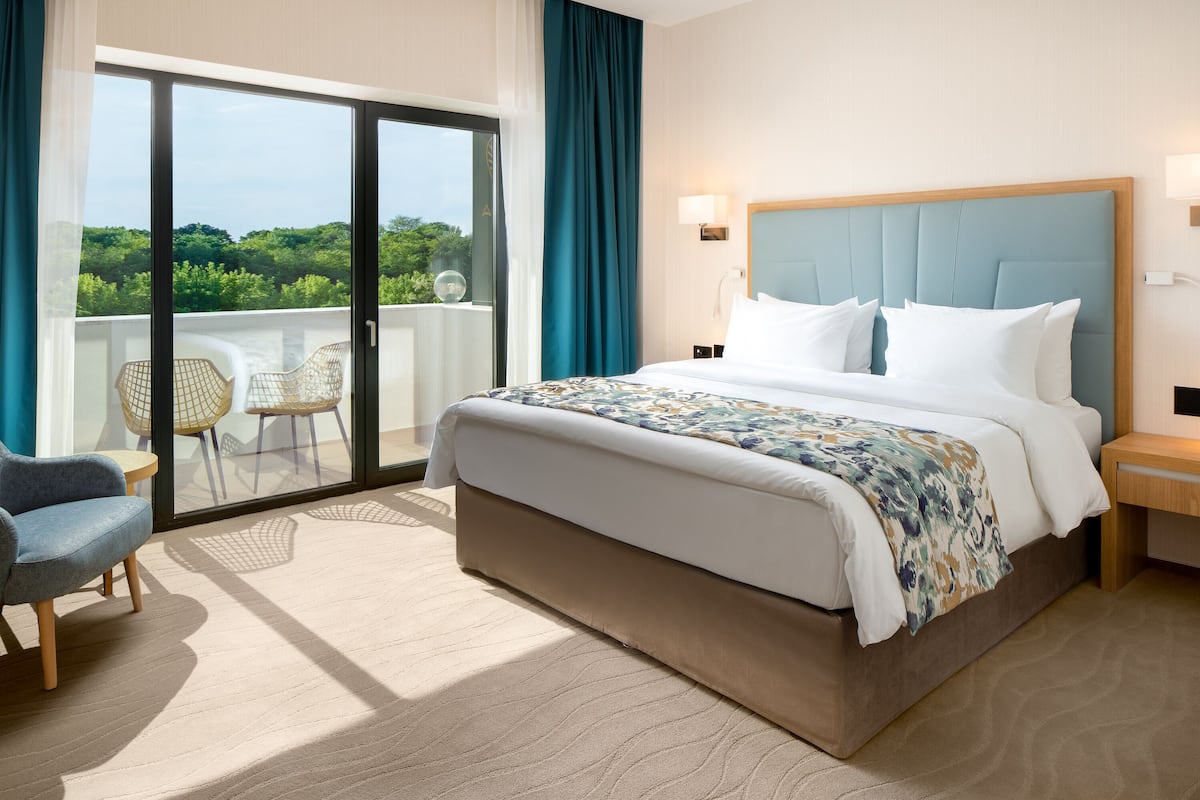 A hotel room with a large bed, a balcony with chairs, and a view of greenery.
