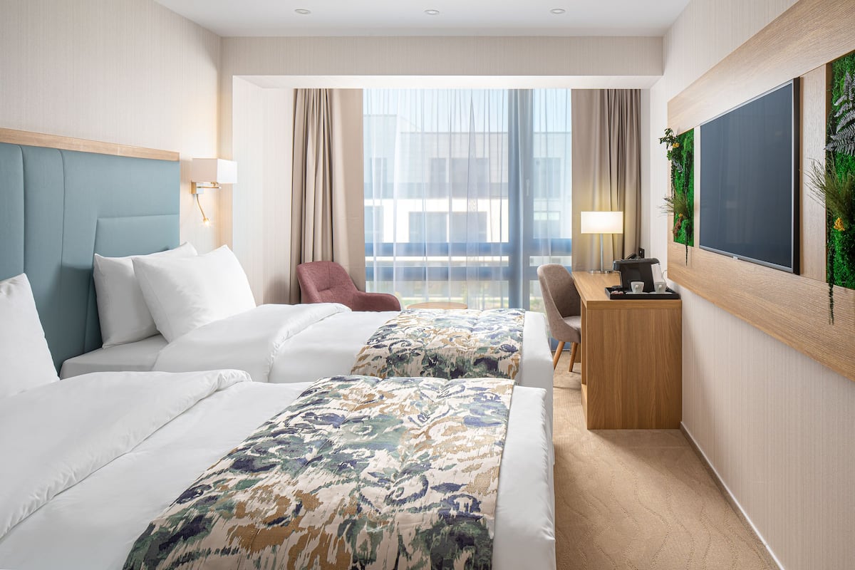 A hotel room with two beds, a desk, a chair, and a large window with curtains.