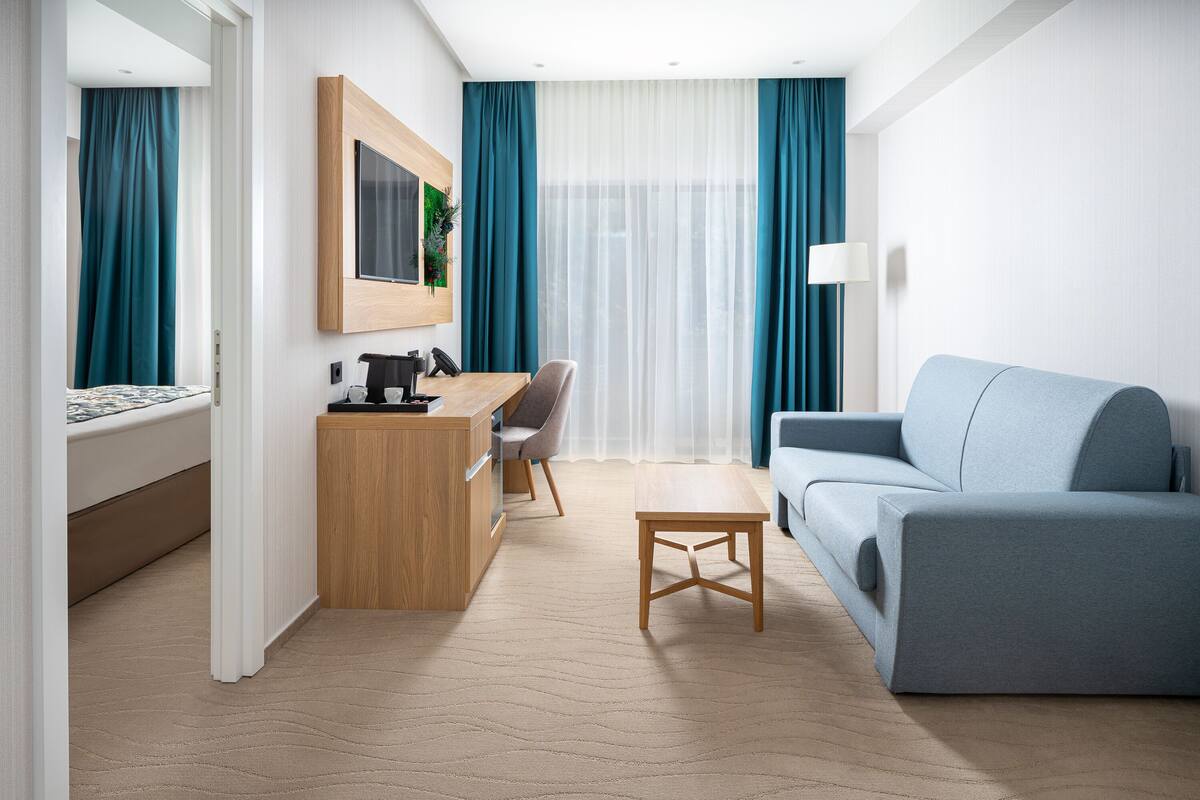 A modern hotel room with a bed, a desk, a chair, a sofa, and a large window with curtains.