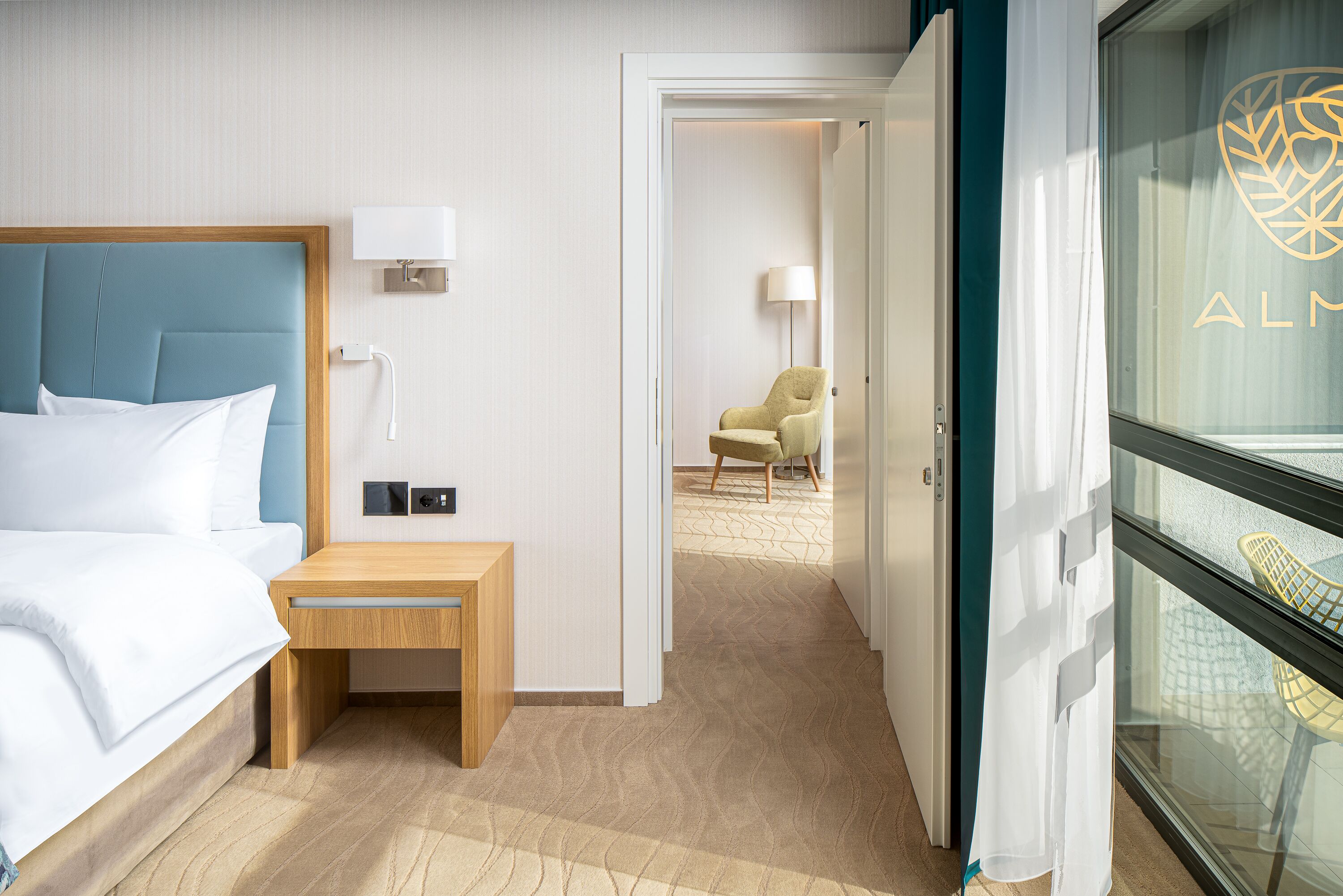 A hotel room with a bed, bedside table, lamp, and a view of another room through an open door.