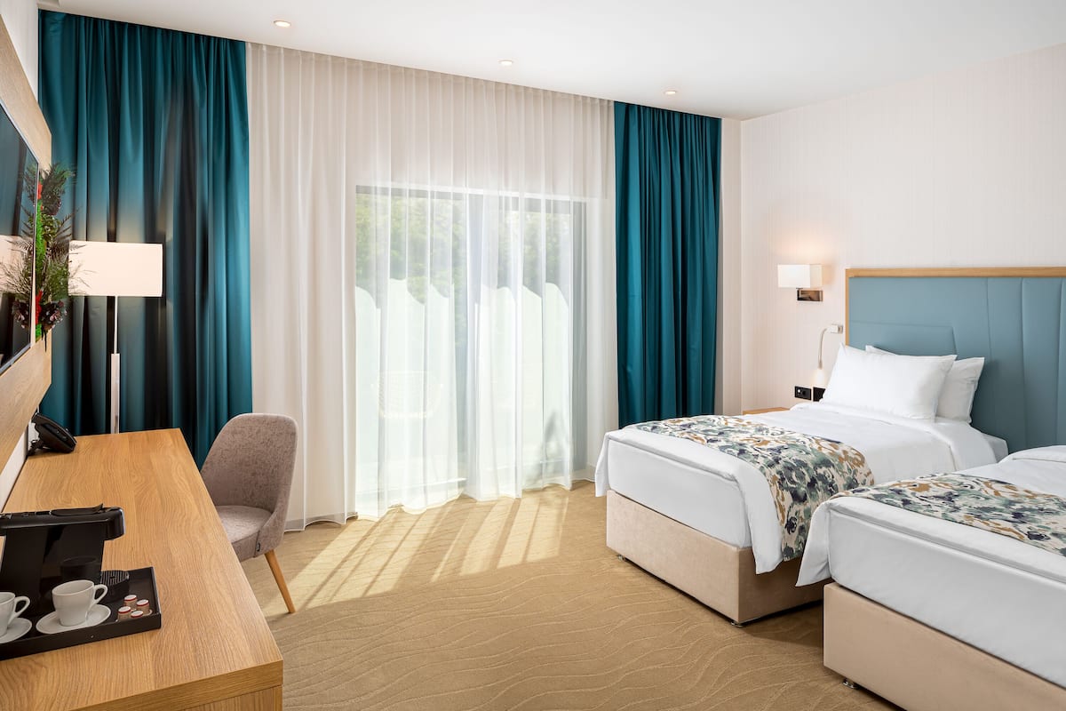 A hotel room with two beds, a desk with a coffee maker, a chair, and a large window with curtains.