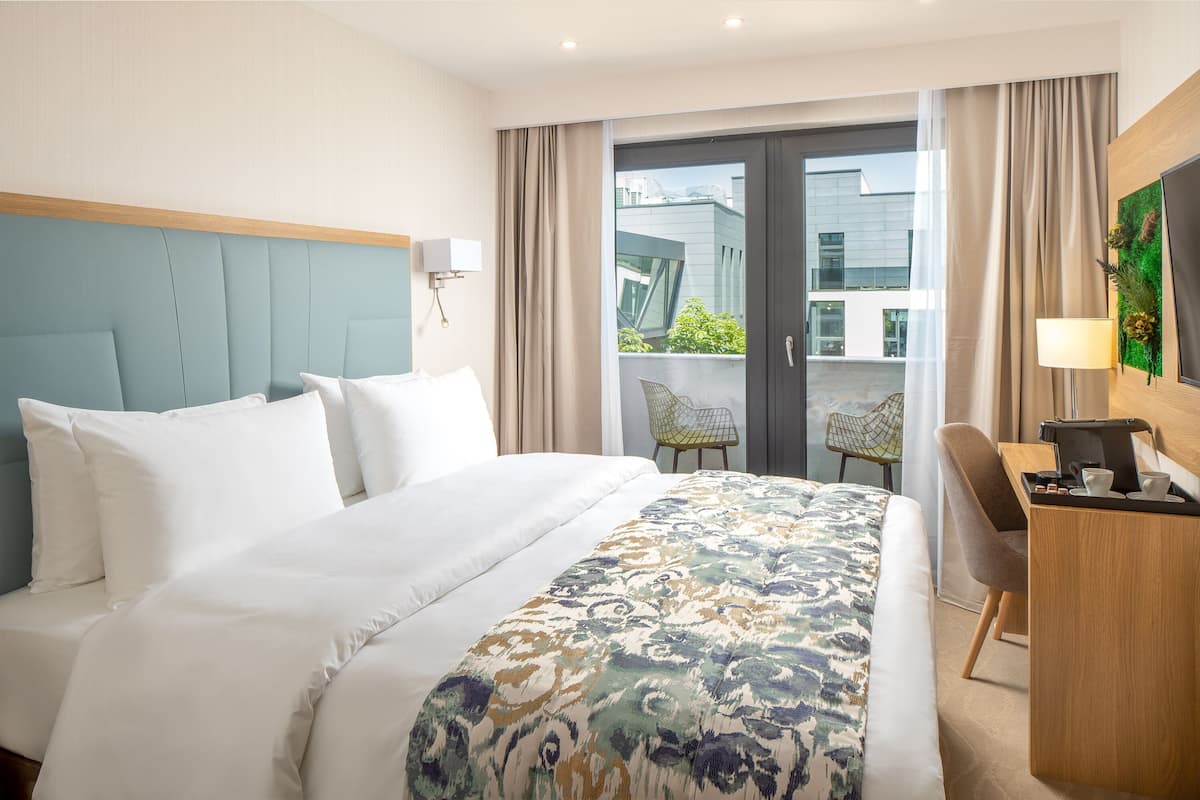 A hotel room with a bed, a desk, a chair, a TV, and a balcony with a view of buildings.