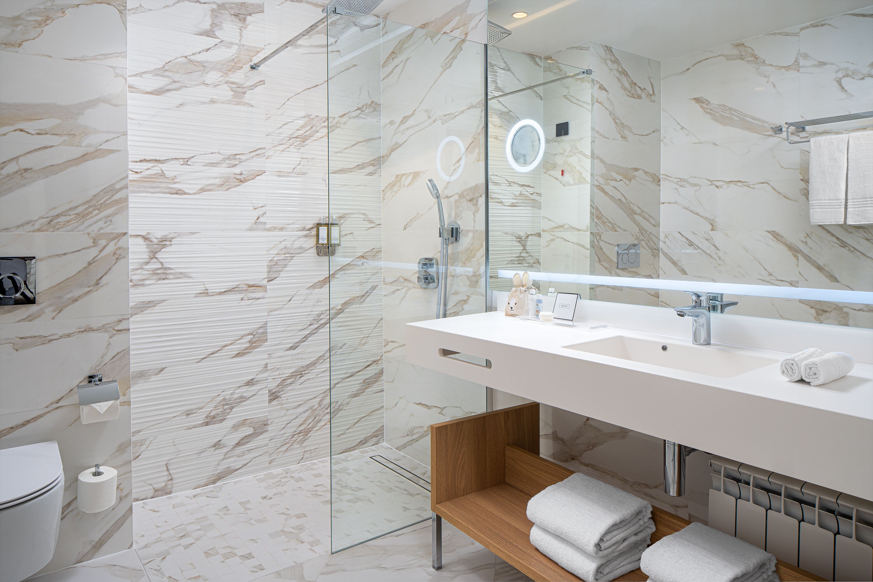 Une salle de bain moderne avec une douche à parois de verre, un grand miroir et une étagère en bois où sont posées des serviettes.