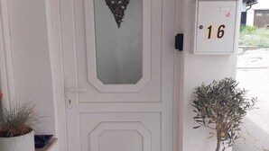 Una puerta blanca con una decoración en forma de corazón, un buzón con el número 16, y una pequeña planta en maceta en el porche.