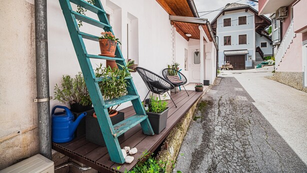 Un estrecho pasillo exterior con cubierta de madera, una escalera color turquesa, plantas en macetas y una silla negra.