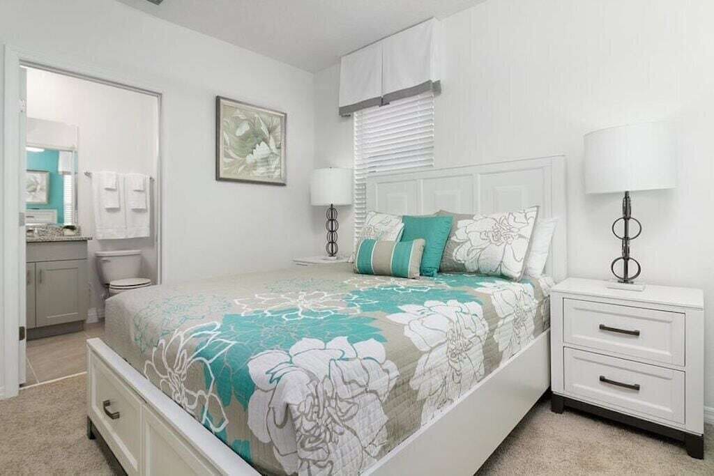 Een slaapkamer met een bed, nachtkastjes, een dressoir, een raam met jaloezieën en een zichtbare badkamer op de achtergrond.