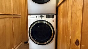 A compact laundry room with a stacked washer and dryer, wooden cabinets, and a small mat on the floor. - Drive up & park 6 ft from this clean, equipped houseboat in Cypress Cove Marina (Buras)