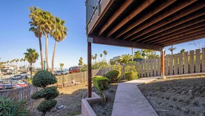 Un espacio al aire libre techado con un sendero de concreto que conduce a una terraza de madera, rodeado de palmeras y una valla de madera.