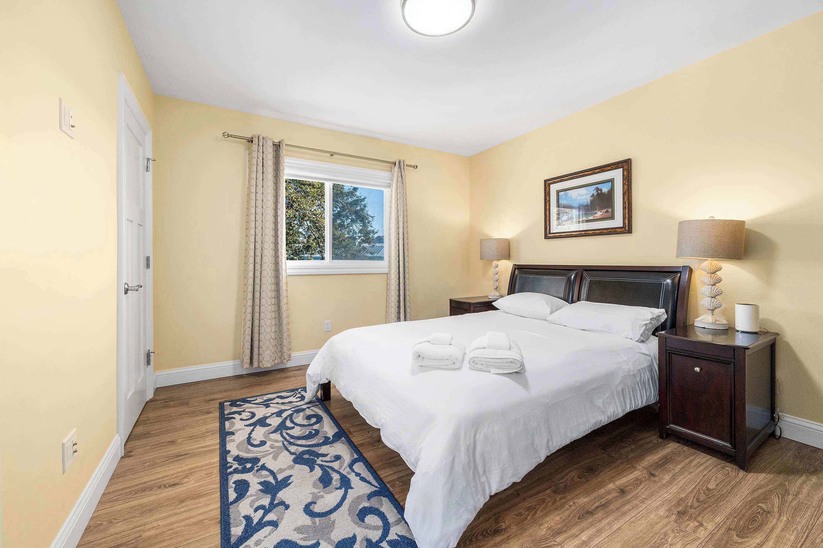 A hotel room with a large bed, two bedside tables with lamps, a window with curtains, a framed picture on the wall, and a blue patterned rug.