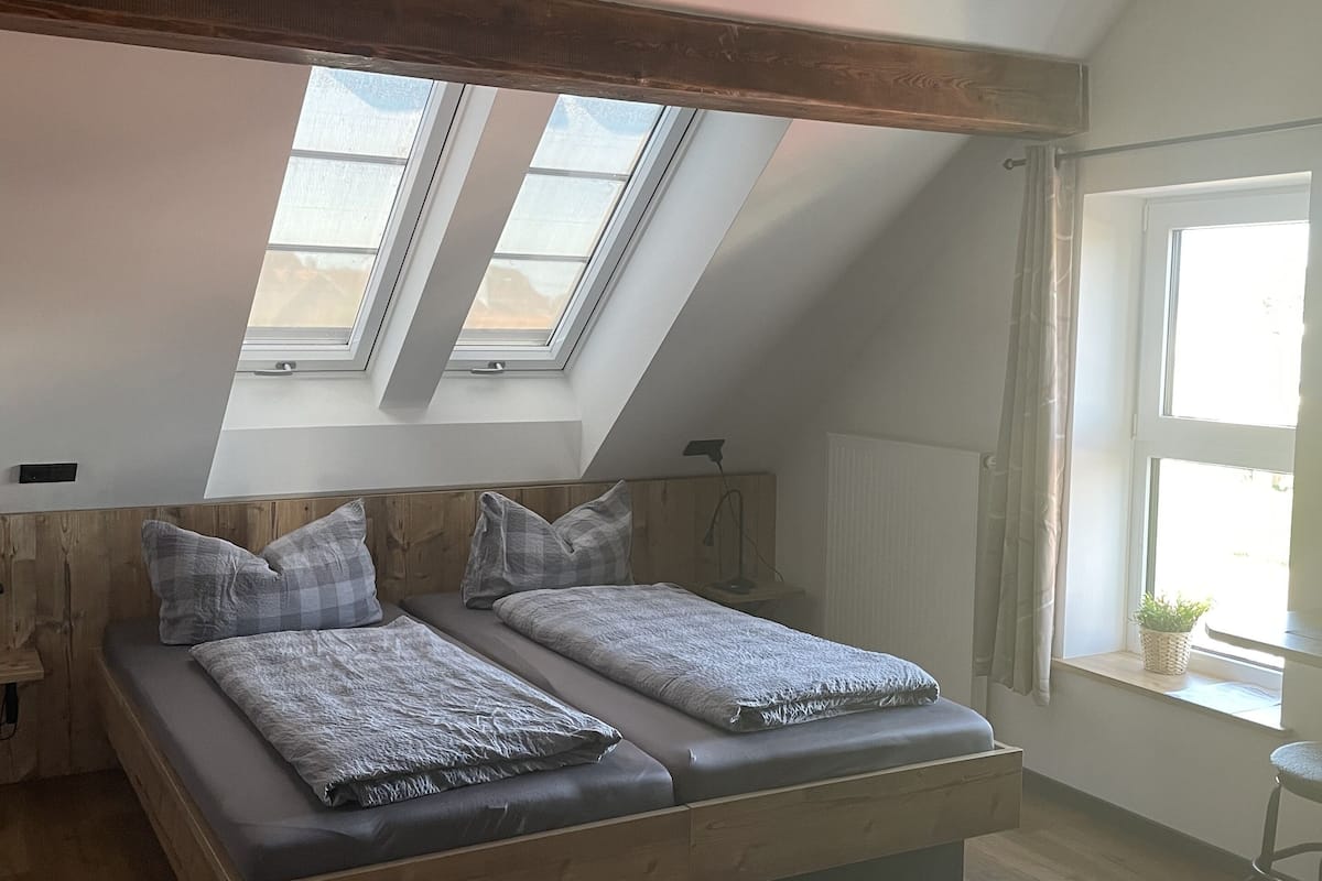 A bedroom with a sloped ceiling, skylights, a bed with grey bedding, wooden headboard, and a small bedside table with a lamp.