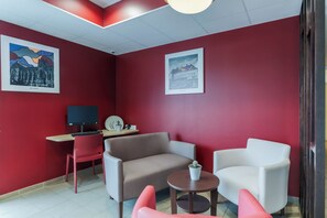 Ein Zimmer mit roten Wänden, einem Schreibtisch mit Computer, zwei Sofas, einem kleinen Tisch mit Pflanze und zwei gerahmten Bildern an der Wand.