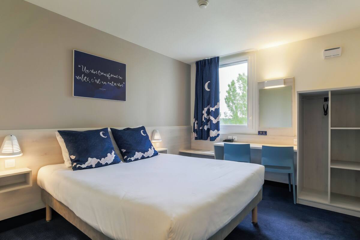 Une chambre d’hôtel comprenant un lit, des tables de chevet, un bureau, une chaise, une armoire et une fenêtre avec des rideaux.