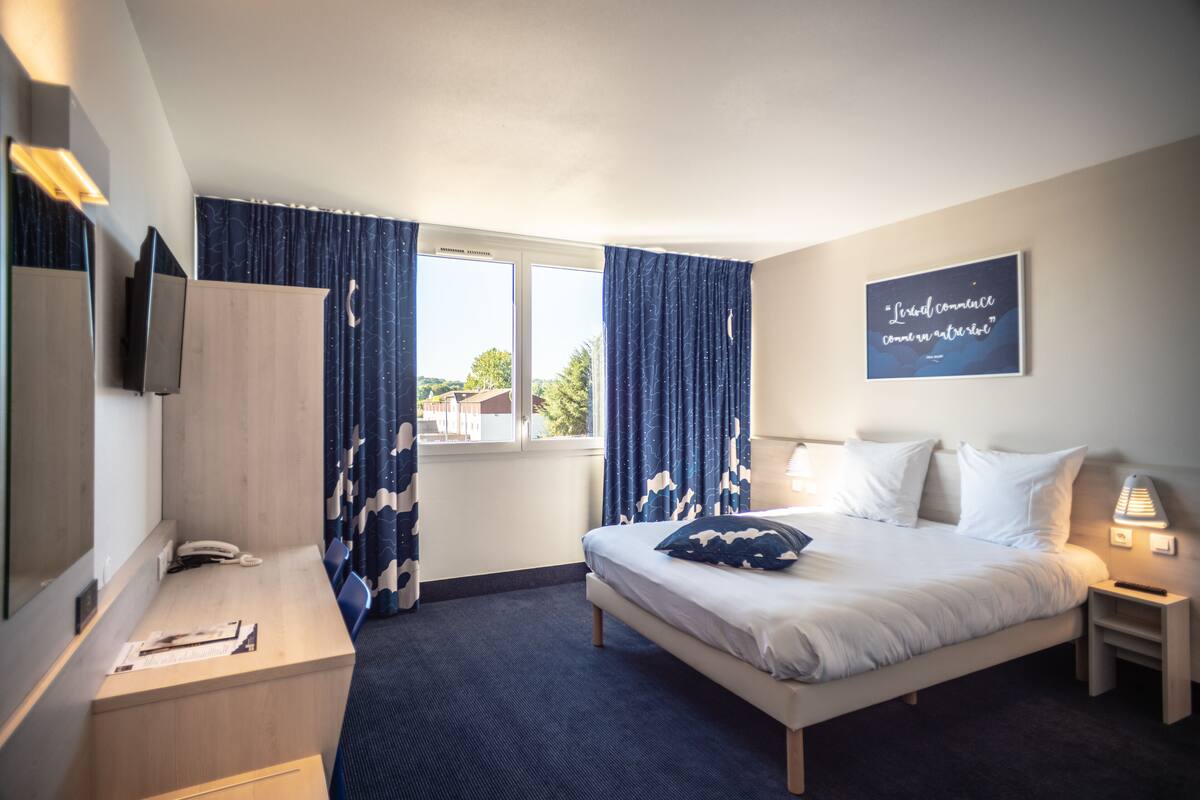 Une chambre d’hôtel comprenant un lit, un bureau, une chaise, une télévision, une fenêtre avec des rideaux et un mur orné d’un cadre contenant une citation.