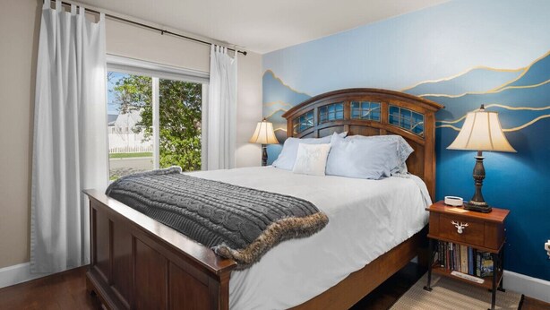 Une chambre avec un grand lit, deux lampes de chevet, une table de chevet avec des livres, une fenêtre avec des rideaux et un mur bleu orné d’une fresque.
