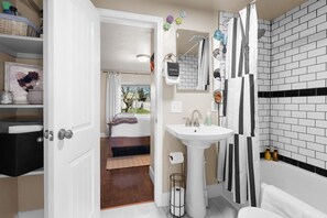Ein Badezimmer mit einem weißen Waschbecken, einem Spiegel und einer Dusche mit schwarz-weißen Streifen. Eine Tür führt zu einem Schlafzimmer mit einem Bett und einem Fenster.