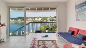 Ein Balkon mit Blick auf ein Gewässer und Gebäude, ein weißer Tisch, ein rotes Sofa und eine weiße Kommode.