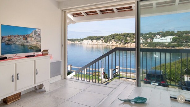 Ein Balkon mit Blick auf eine Küstenlandschaft, eine weiße Kommode mit einem Gemälde und ein kleiner Tisch mit einem dekorativen Gegenstand.