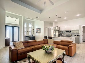 A spacious living area with a brown leather sectional sofa, a wooden coffee table with a floral centerpiece, and a kitchen in the background with modern appliances.