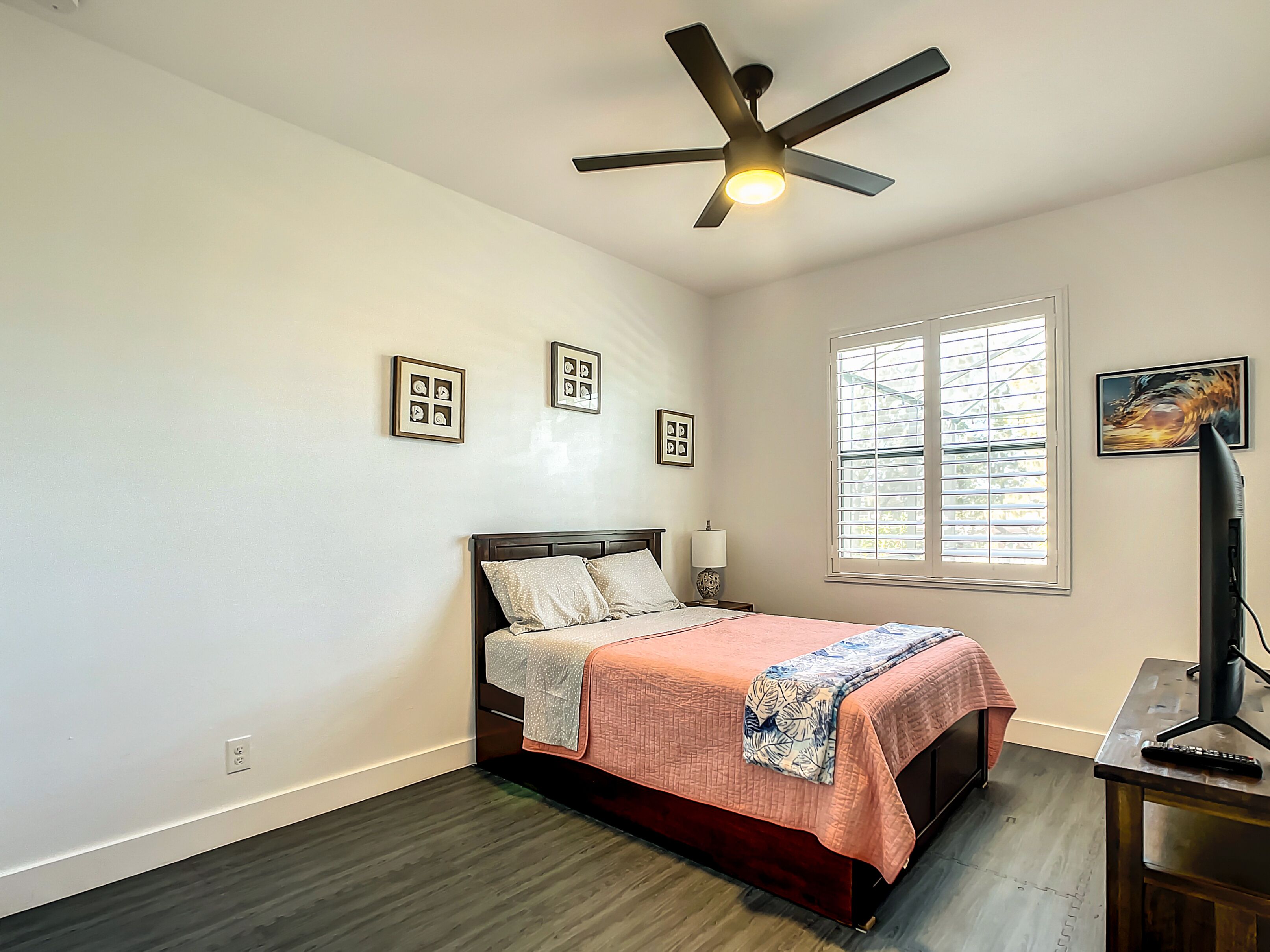 Un dormitorio con cama, ventilador de techo, ventana con persianas y escritorio con lámpara.