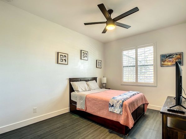 A bedroom with a bed, a ceiling fan, a window with blinds, and a desk with a lamp.