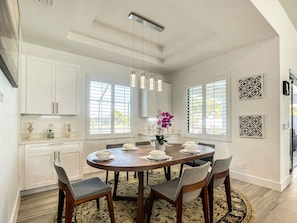 A dining area with a round table, chairs, and a vase of flowers. There are windows with blinds, a ceiling light fixture, and wall-mounted decorative panels.