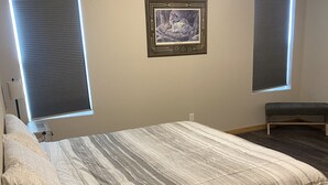 A bedroom with a bed, two windows with blinds, a wall-mounted picture, and a bench. - Brand New Duplex Across the road from Devils Lake w/lake access (Warwick)
