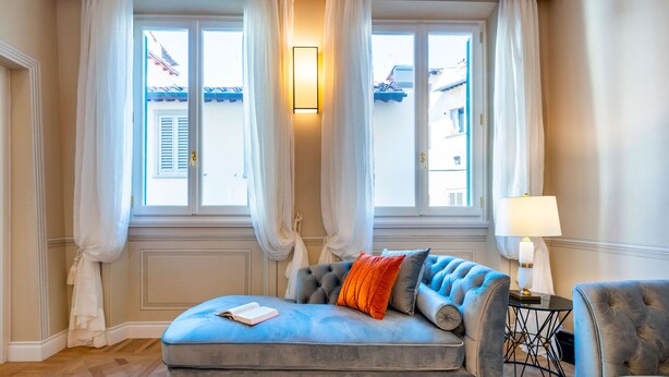 Ein Zimmer mit einem blauen Sofa, einem kleinen Tisch mit einer Lampe und Blick auf Gebäude durch große Fenster.