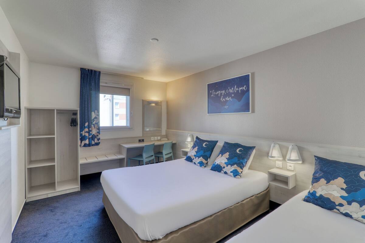 Habitación de hotel con cama, lámparas de noche, escritorio, silla, armario y ventana con cortinas.