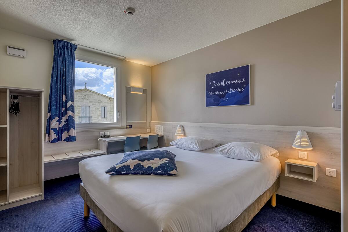 Habitación de hotel con cama, mesitas de noche, ventana con vistas, un banco empotrado y una obra de arte en la pared.