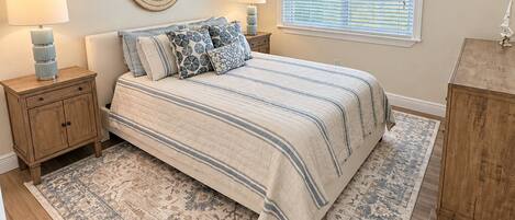 A bedroom with a bed, two bedside tables, a ceiling fan, a window with blinds, and a decorative wall piece.