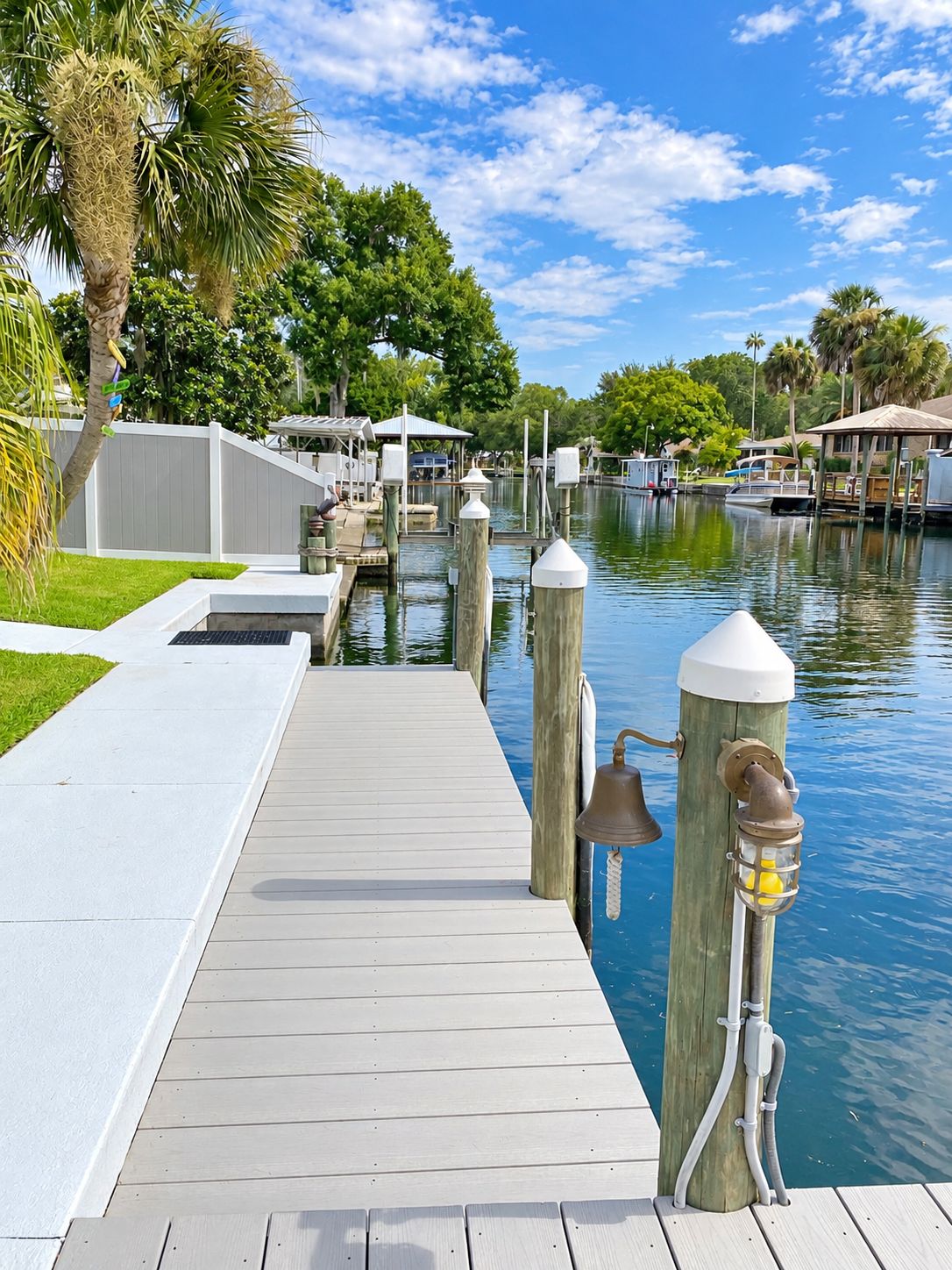 A wooden dock leading to a marina with boats, a white fence, and lush greenery.