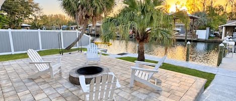 An outdoor patio with a fire pit, Adirondack chairs, and a view of a body of water during sunset.