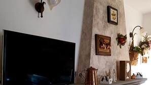 A living room with a flat-screen TV mounted on the wall, a stone fireplace with a mantel, and decorative items including a copper kettle and framed pictures. - Romantic house with swimming pool near Bologna & Florence CIN: IT047018C2OXJNJAB8 (Treppio)