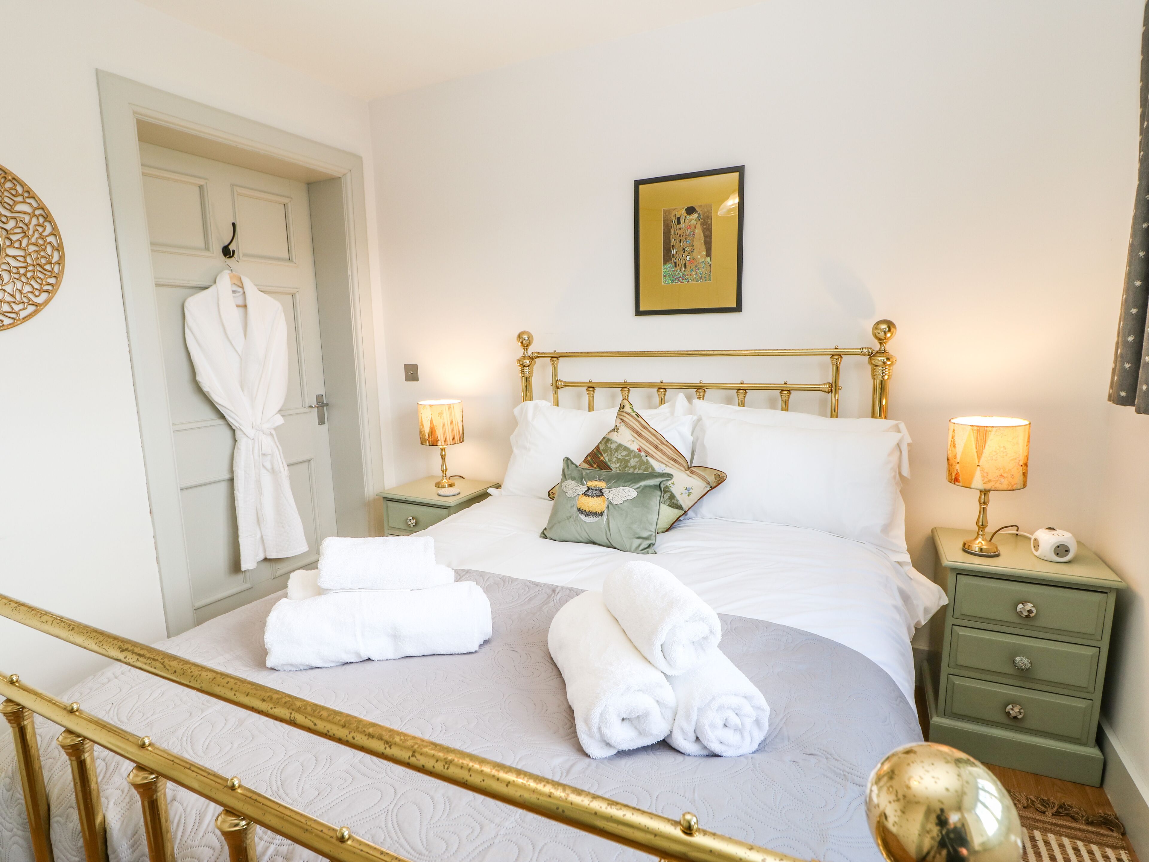 Una habitación de hotel con una cama, dos mesitas de noche, una lámpara de noche, cabecero con marco dorado, una obra de arte enmarcada sobre la cama, una bata colgada en la puerta y un par de toallas sobre la cama.